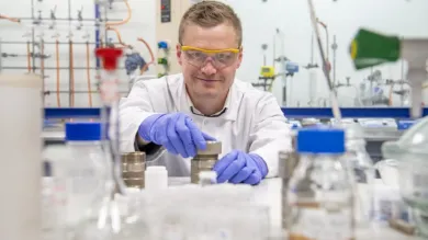 A chemistry researcher in goggles, gloves and white coat works with equipment in the lab