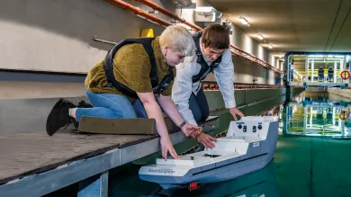 A student crouches down to place a model boat into the water of the university towing tank, a researcher at her side