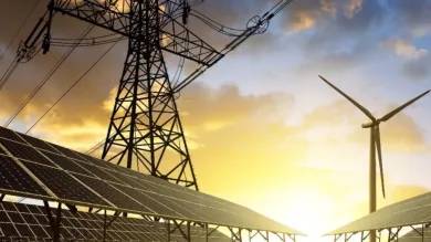 A low-angle view of solar panels, a wind turbine and a pylon, with the sun rising in the background