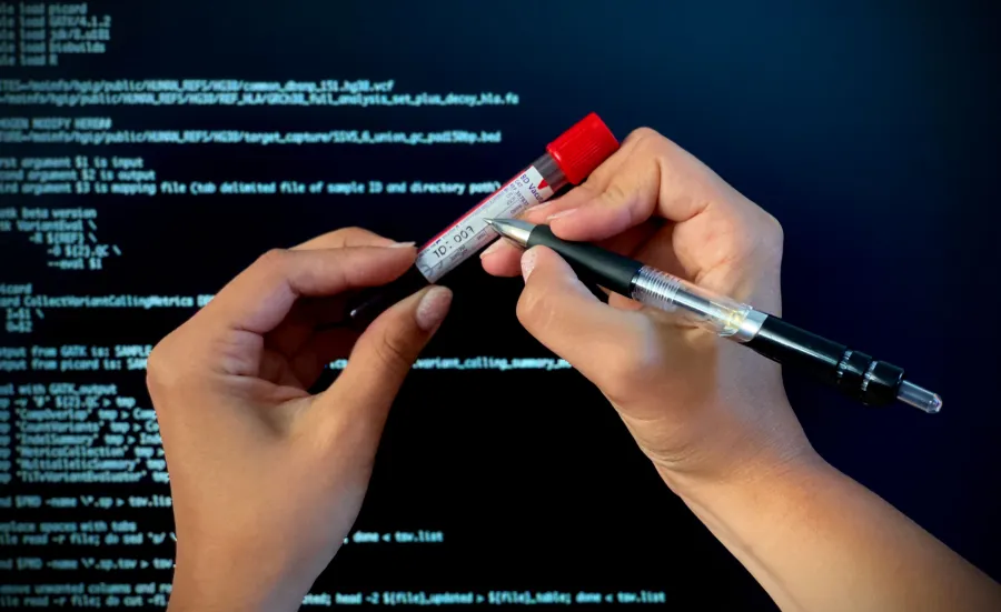 Somebody writes the time and date on a blood sample in a vial, against a background of a screen of data to represent genomic data. 