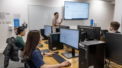Three students in front of monitors in a classroom listen as a lecturer makes a point