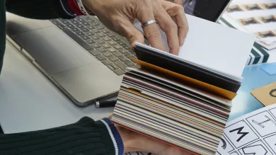 Two hands flicking through a swatch book. Hand drawn sketches in the foreground, with contact sheets and a laptop