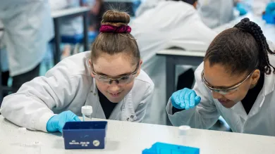 Two school children in lab coats and safety goggles examining laboratory equipment with focus, surrounded by others in a laboratory setting.