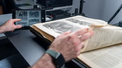 A close-up of a person's hands working with an open historical book on a specialized scanning table. One hand is turning a delicate page with illustrations, while the other holds a tablet displaying a digital scan of the same page. The workspace includes modern scanning equipment and cables in the background, blending traditional and digital technologies.