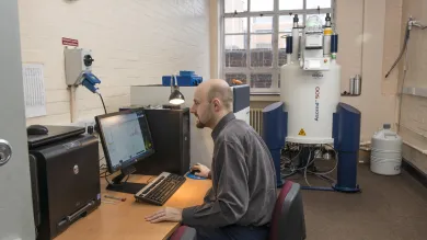 A man working  on a computer in a room with a Nuclear Magnetic Resonance machine in the background