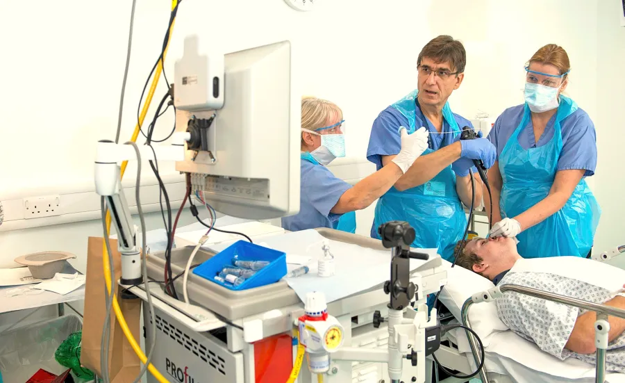 Three medical professionals conduct a procedure on a patient lying on a hospital bed, surrounded by medical equipment and supplies, in a sterile clinical environment.