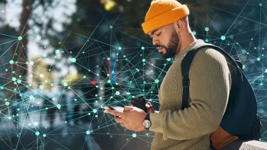 A man in a yellow beanie and olive-green sweater, engrossed in a smartphone, holding a disposable coffee cup, and wearing a backpack. He is standing against a backdrop of an abstract digital network design with interconnected nodes and lines.