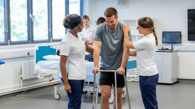 A patient, supported by 2 students, learns to walk using a walking frame.