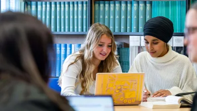 Two students discussing work in a library with a yellow laptop.