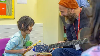 A bearded man wearing an orange knit hat and visitor lanyard plays guitar while interacting with a young child in a light blue shirt who is holding drumsticks. They appear to be in a colorful classroom or community center setting, engaged in a musical activity together.