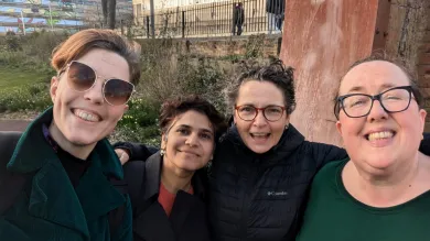 Four women pose together for a cheerful group selfie outdoors, all smiling warmly at the camera. They are dressed in winter clothing including coats and jackets, with urban buildings and greenery visible in the background.