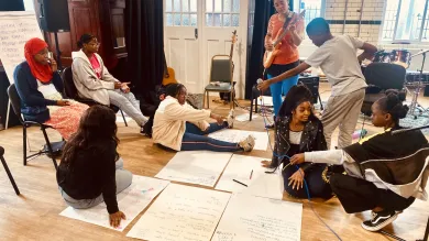 A group of young people participate in a creative workshop or music session in a large room with wooden floors, surrounded by large sheets of paper with writing and musical instruments including guitars and drums. Some participants are seated on chairs while others sit on the floor, with one person playing guitar and recording equipment visible in the scene.Retry