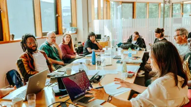 A diverse group of people sit around a large wooden conference table in a bright, modern meeting room with floor-to-ceiling windows, engaged in discussion with laptops, notebooks, and coffee cups spread across the table. The natural lighting and contemporary architecture create a collaborative workspace atmosphere for what appears to be a business meeting or workshop.