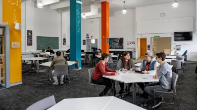 A study and social space on a university campus. In the foreground, there is a group of four students sat around a table. There are more tables with students in the background, and the pillars around the room have mathematical equations written on them.