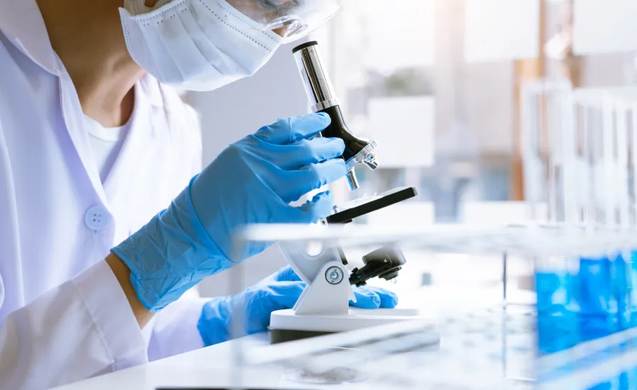 A person in a lab coat using a microscope, wearing a face mask and blue gloves.