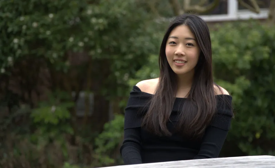 Student sitting outdoors with green shrubs behind her. She is smiling at the camera.