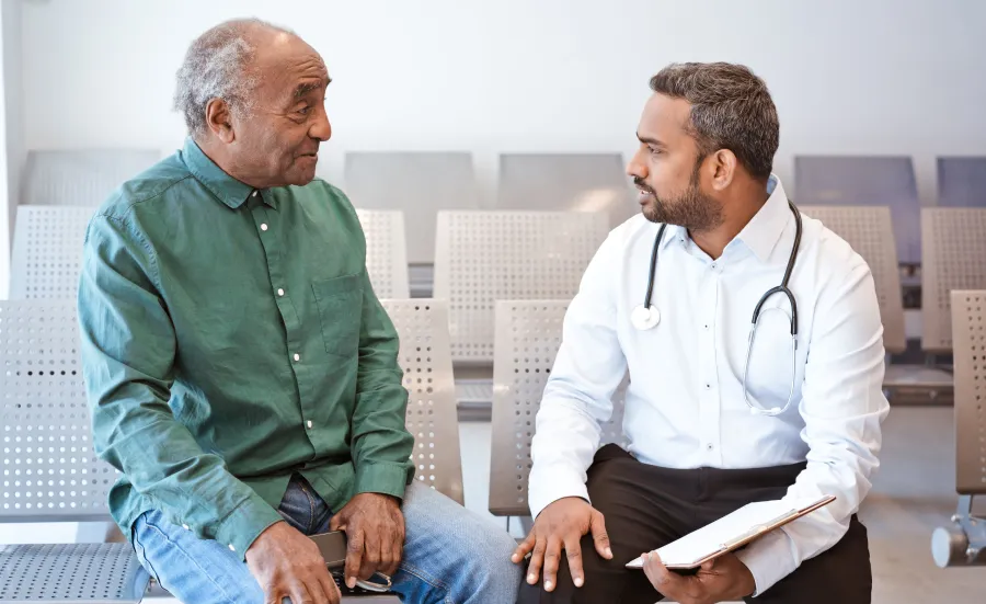 A doctor consulting a patient in a hospital waiting room. The doctor is holding a clipboard and has a stethoscope around their neck.