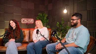 Three people sat on chairs and a sofa, in front of microphones in a podcast recording studio.