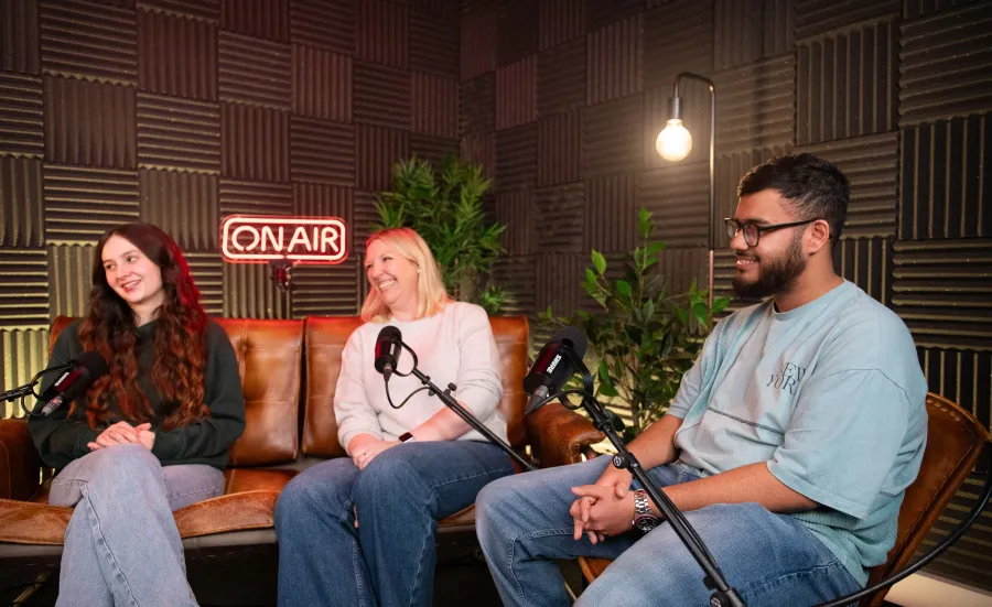 Three people sat on chairs and a sofa, in front of microphones in a podcast recording studio.