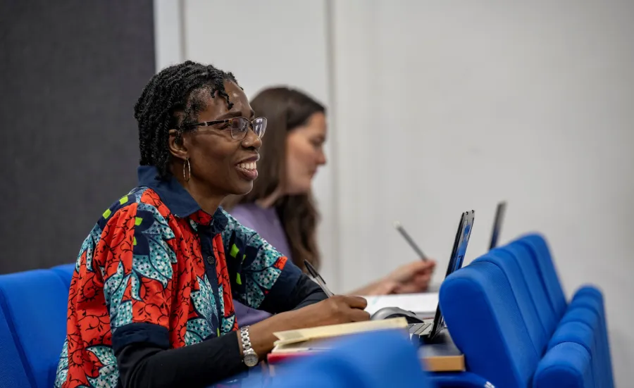 Ogene Adoh sat in a lecture classroom. She has a laptop and notebook in front of her and is looking towards the front of the classroom. 