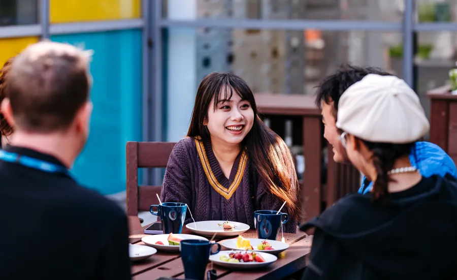 Students sitting outside at a table chatting. There are plates of food and mugs on the table.