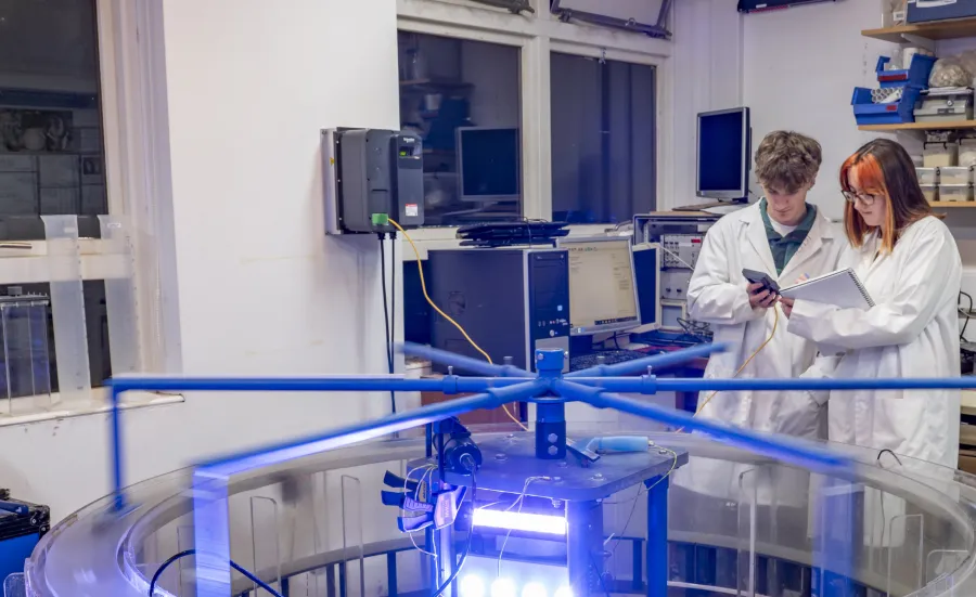 Students in white coats recording data from a rotating blue mechanical arm above a large circular transparent tank filled with liquid.