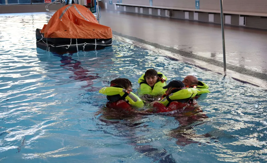 Four people wearing bright yellow life jackets and red immersion suits are floating in a swimming pool, huddled together in a survival position. 