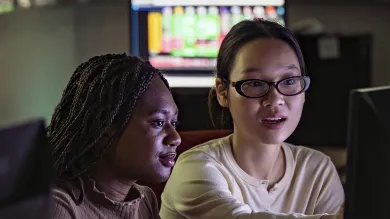 Two students seated together in the Bloomberg suite. One student gestures towards something on a computer screen.