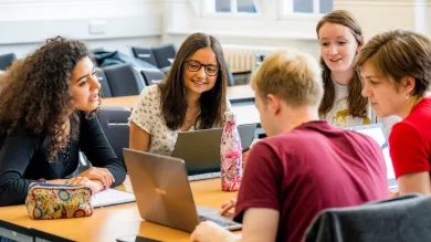 Five students gathered around a table using laptops and discussing work together.