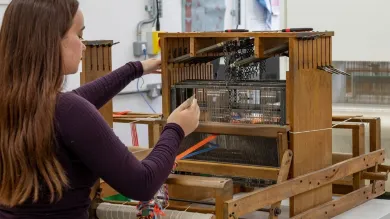 Student adjusting a wooden weaving loom in a textile studio, with threads and tools in the foreground and a modern loom and colourful yarns in the background.