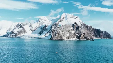Snow-covered Antarctic island rising from clear blue ocean beneath a bright sky, with a small boat visible in the distance.