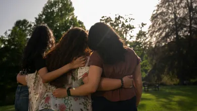 Three international students put their arms around each other and look away from the camera, towards a horizon lined with lush green trees and bright sunny skies