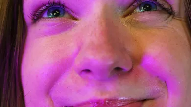 The camera leans close to the face of a student bathed in a warm purple light. She looks up, smiling expectantly