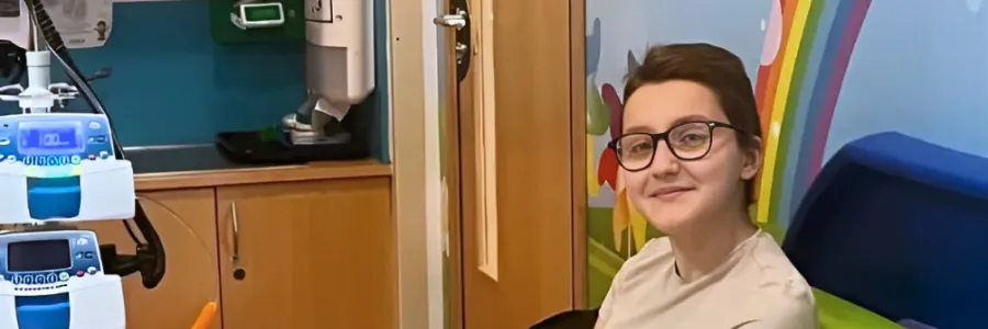 Gio sitting on a colourful bench in a hospital room, reading a book and smiling, with a medical device and snacks nearby.