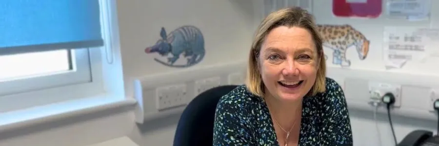 Professor Juliet Gray sitting at her desk, smiling. There are pangolin and leopard stickers on a white wall in the background. The window behind her is mostly covered by a light blue blind.