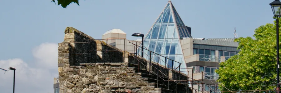 View of a historic stone wall with staircase and metal railings in the foreground, juxtaposed against a modern building featuring a glass pyramid roof. The scene includes leafy tree branches above and a traditional street lamp on the right, highlighting the contrast between medieval and contemporary architecture in an urban setting.