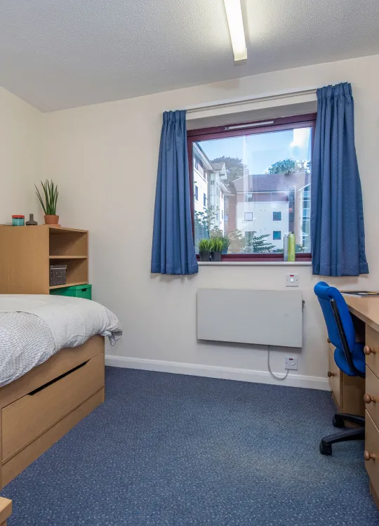 A window lets light into a bedroom where a neatly made bed and desk can be seen.
