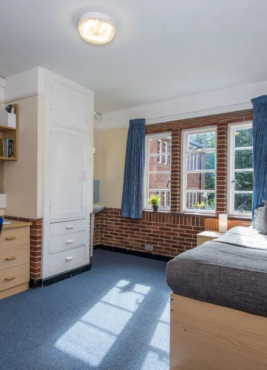 Light from a large window fills a spacious bedroom with desk, chair and neatly made bed.