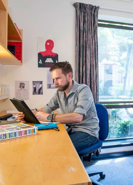 A student sits looking at a tablet computer while working at a desk. A bed and wardrobe can be seen behind him, an open window lets light into the room.