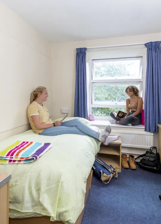 Two students chatting in a bedroom. One sits browsing with her tablet on a single bed while the other looks up from their book, sat on the windowsill.
