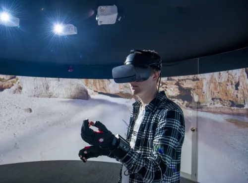 A student wearing a virtual reality headset and gloves, standing in front of a 360 degree video wall displaying what he can see.