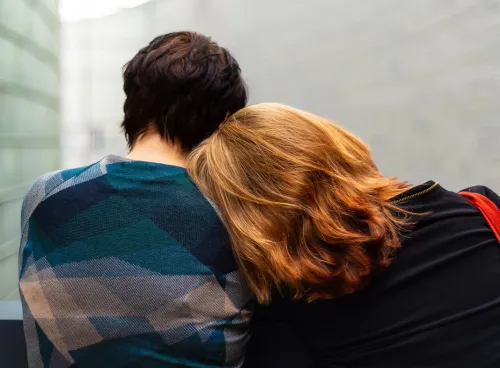 Two people sitting down, facing away from us. One is leaning into the other.