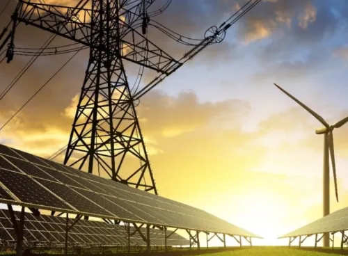 A low-angle view of solar panels, a wind turbine and a pylon, with the sun rising in the background