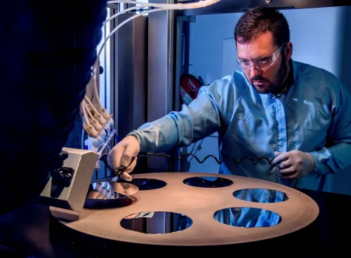 A bearded young scientist in protective overalls and glasses works with a flame hydrolysis deposition (FHD) system