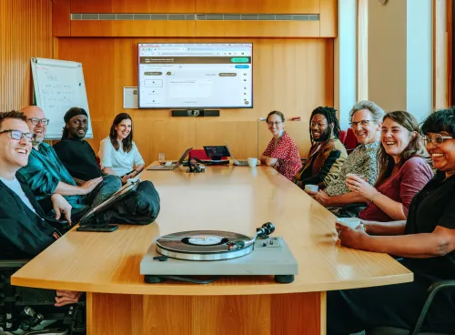  A group of nine people is seated around a wooden conference table, smiling and engaged in a meeting. Laptops are open, and a turntable with a vinyl record sits at the front of the table. A large screen behind them shows a web interface, and a whiteboard with notes is visible. The mood is friendly and collaborative in a modern, well-lit room.
