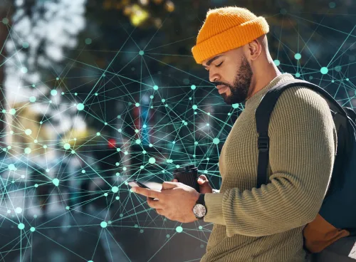 A man in a yellow beanie and olive-green sweater, engrossed in a smartphone, holding a disposable coffee cup, and wearing a backpack. He is standing against a backdrop of an abstract digital network design with interconnected nodes and lines.