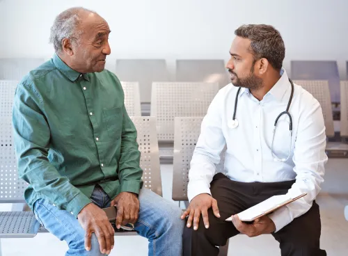 A doctor consulting a patient in a hospital waiting room. The doctor is holding a clipboard and has a stethoscope around their neck.