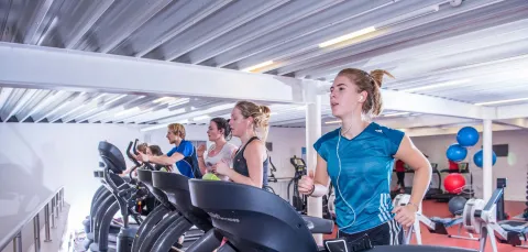 Group of students in gym, running on treadmills surrounded by rowing machines and other gym equipment.