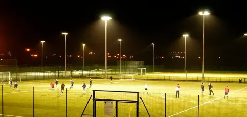Wide view of floodlit pitches on a dark evening. A group of people are playing football.