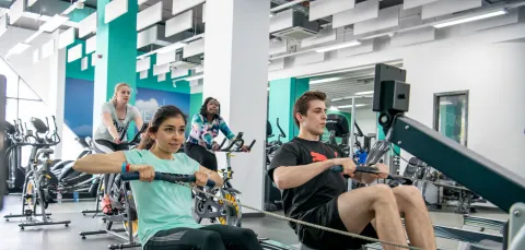 Group of students working out on rowing and cycling machines in a bright, modern gym.